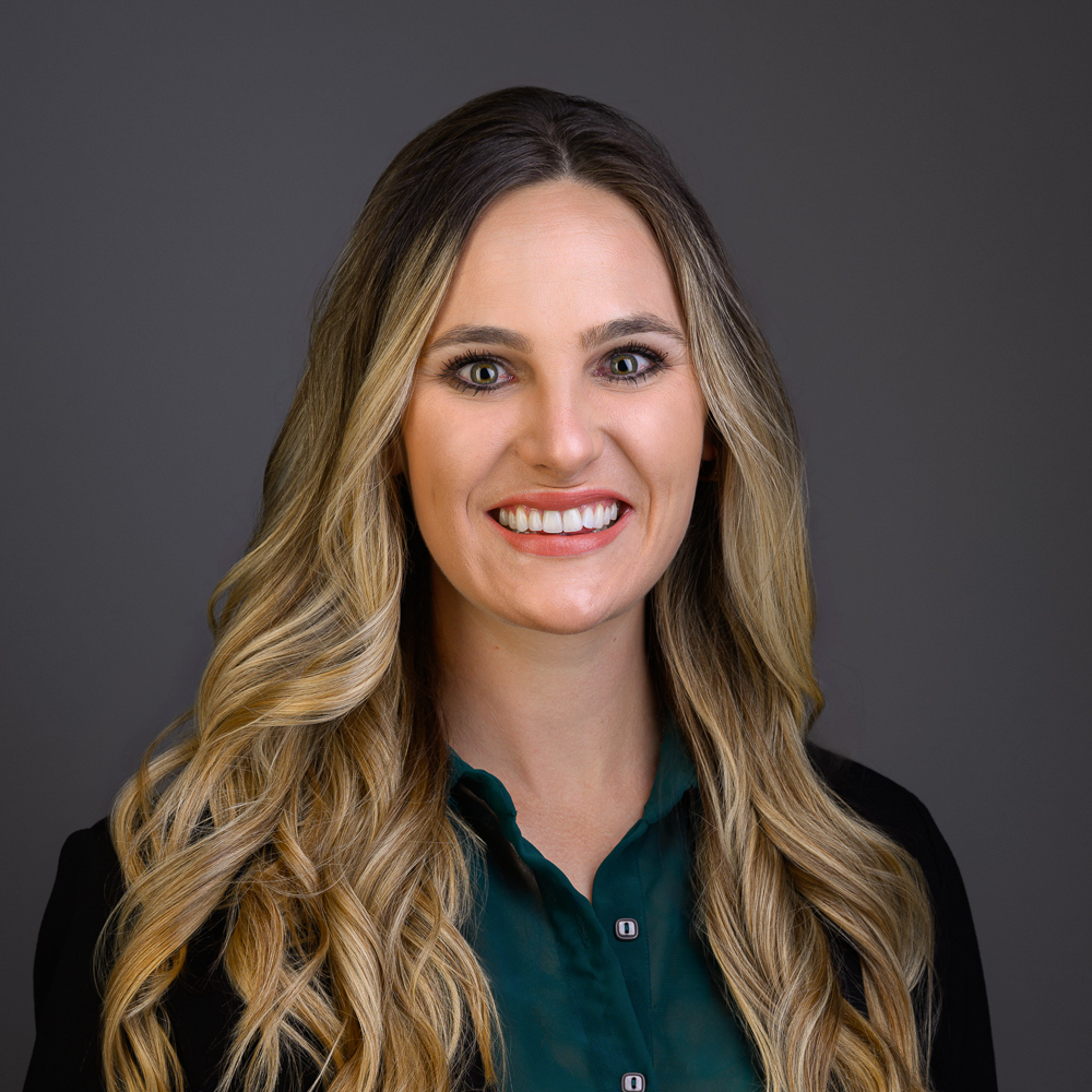 Professional headshot of smiling woman with long wavy brown hair wearing dark blazer and teal blouse against gray background