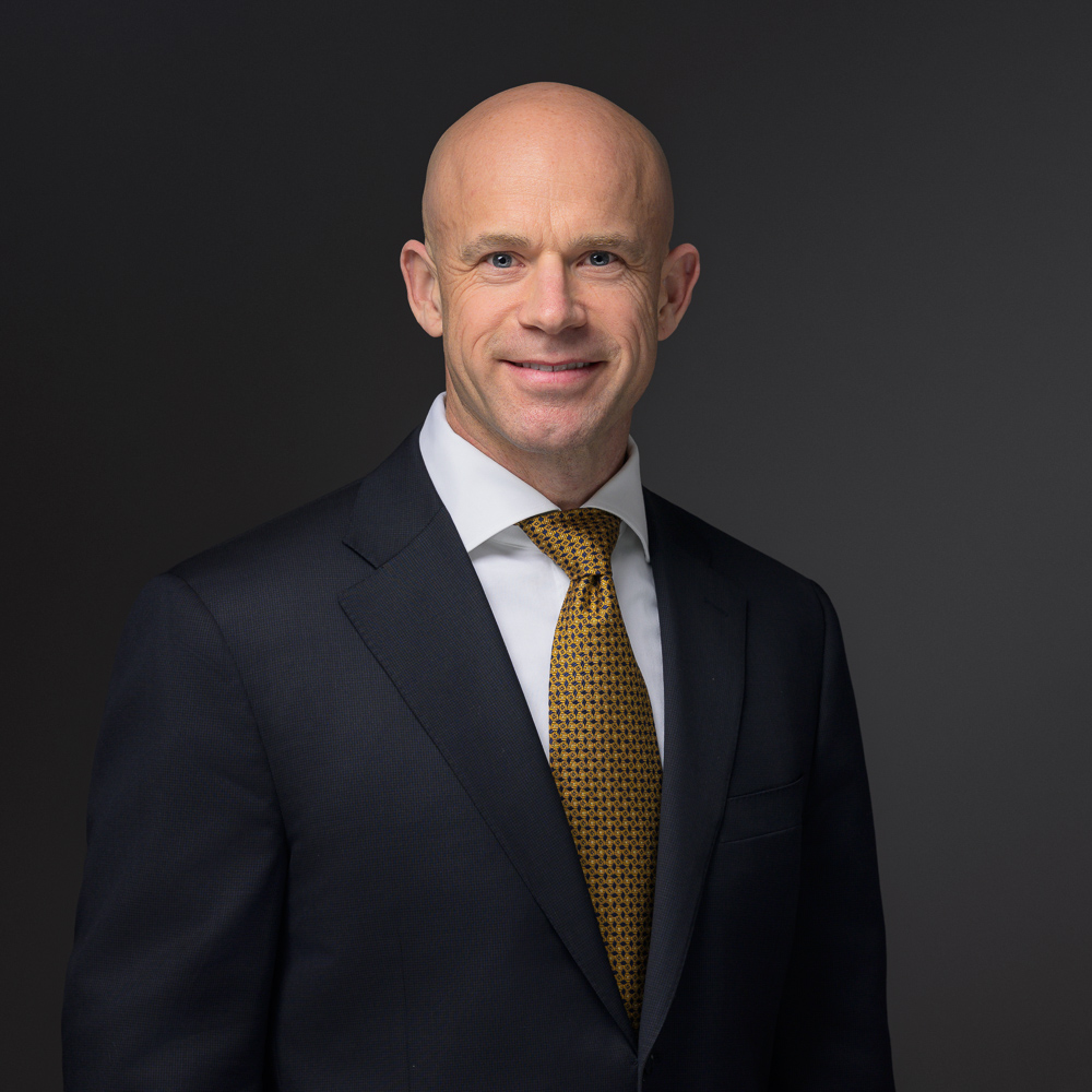 Professional man in dark suit and gold patterned tie smiling at camera against dark background