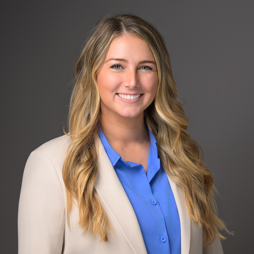 Professional headshot of a smiling woman with long blonde hair wearing a blue blouse and cream cardigan against a gray background