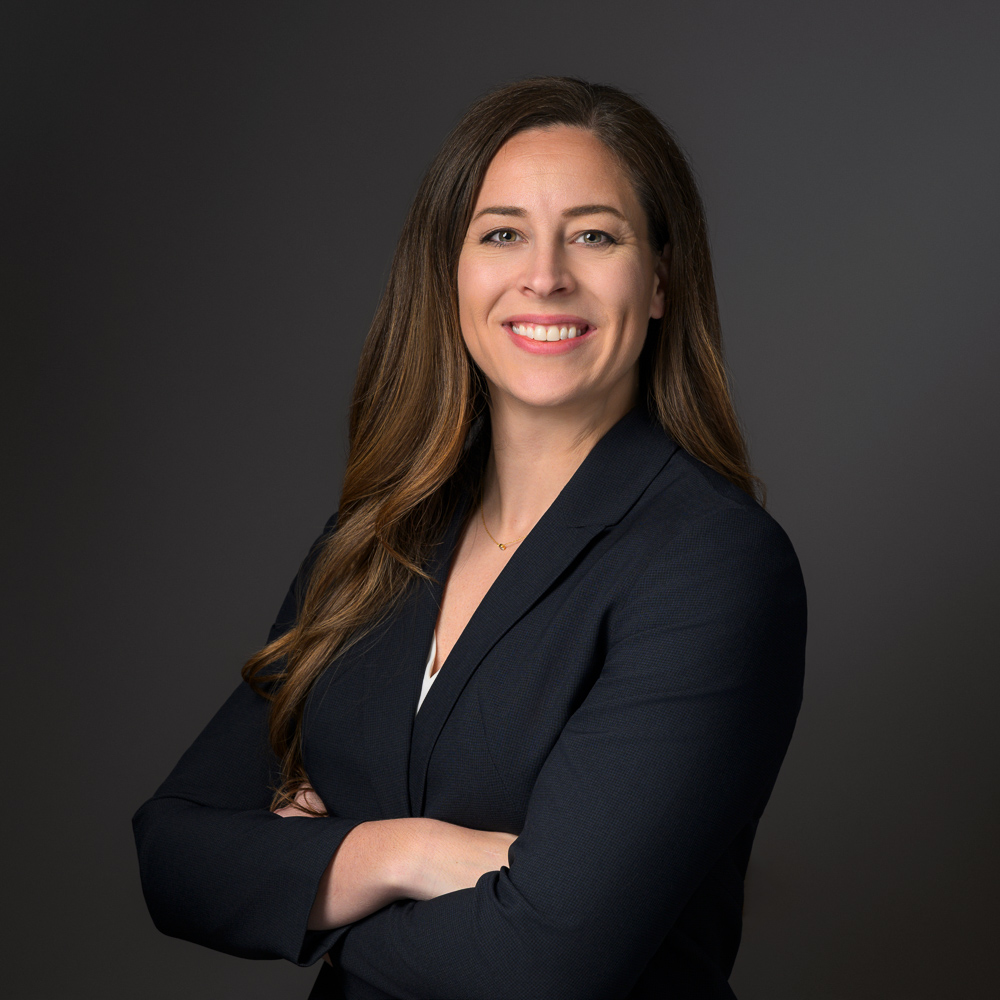 Professional woman with long brown hair smiling at camera, wearing dark blazer against gray background