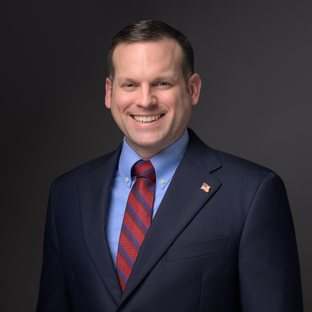Professional headshot of a man in a dark blazer, light blue shirt, and red striped tie, smiling at the camera against a gray background