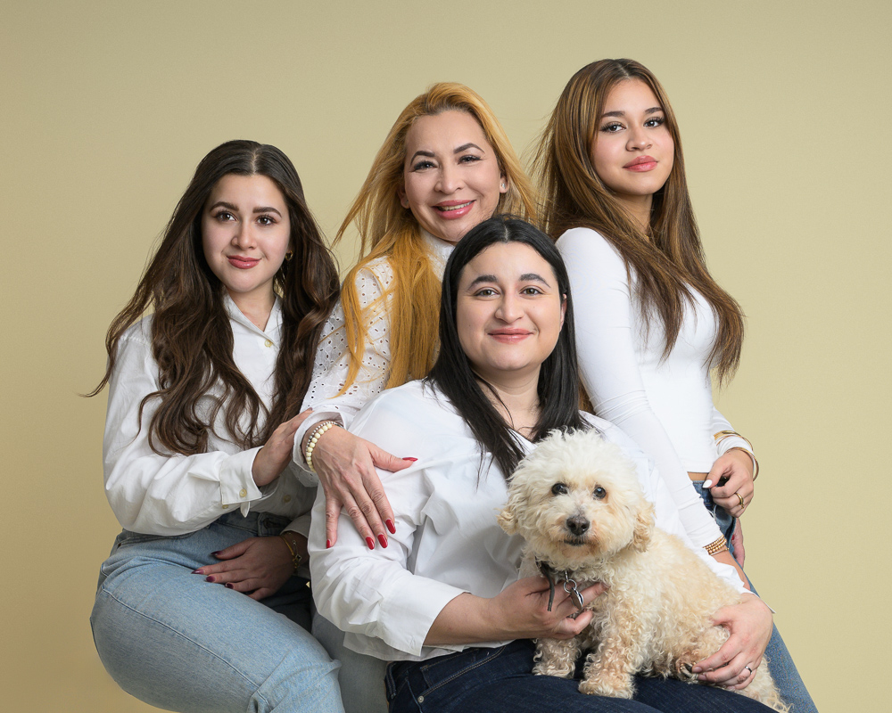 Four women and a cream-colored dog pose together against a beige background for a family portrait