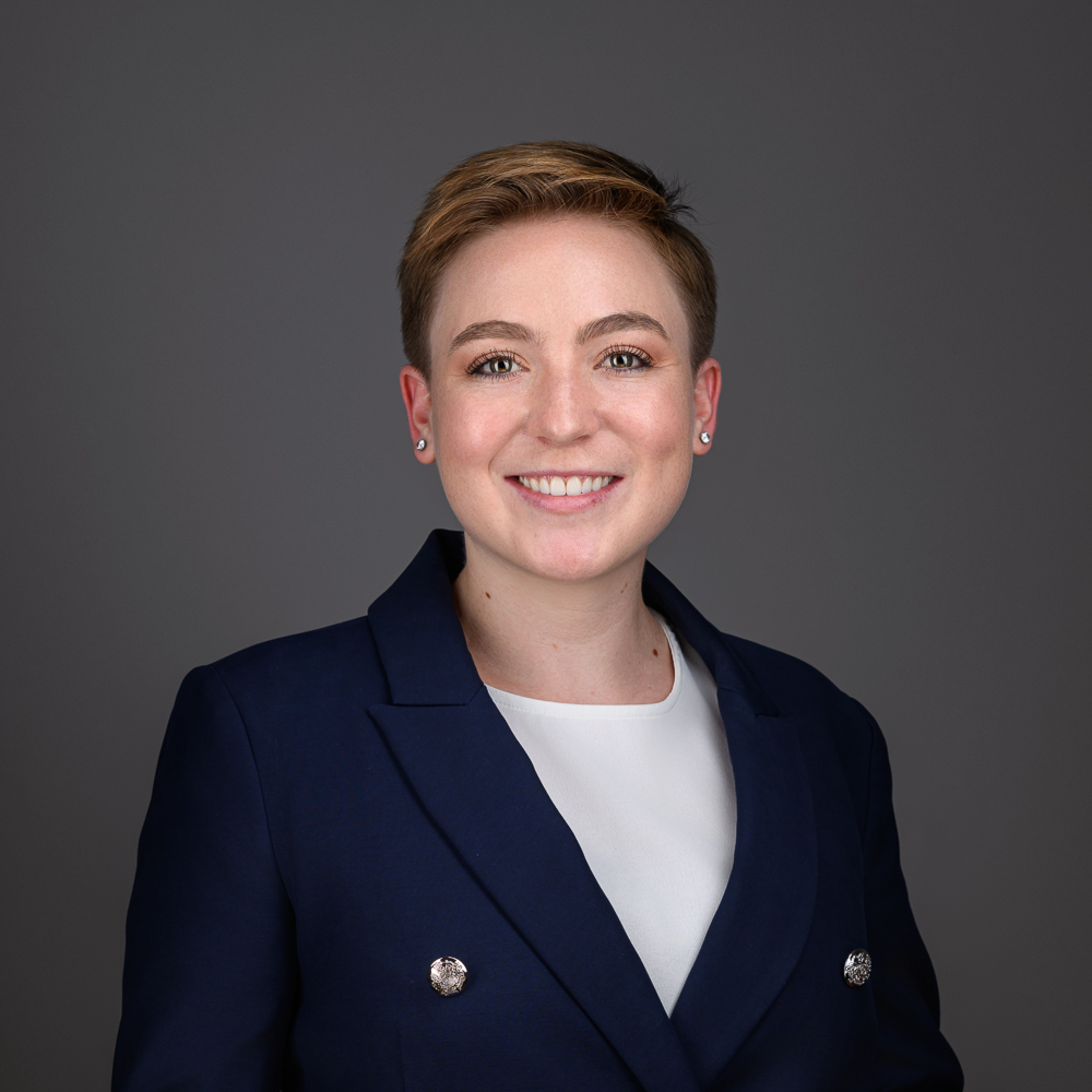 Professional headshot of a smiling woman with short reddish-brown hair wearing a dark navy blazer over a white shirt