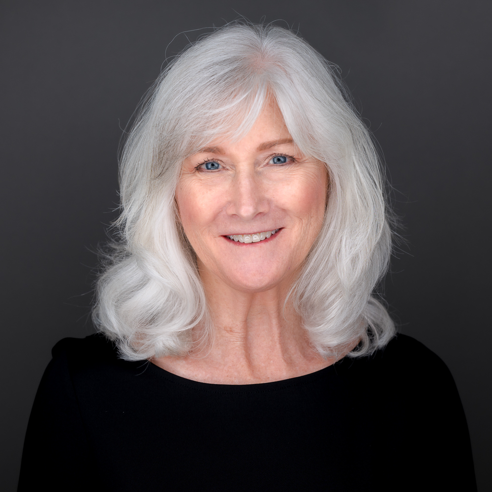 Professional headshot of a woman with shoulder-length white hair and blue eyes, smiling at the camera against a gray background